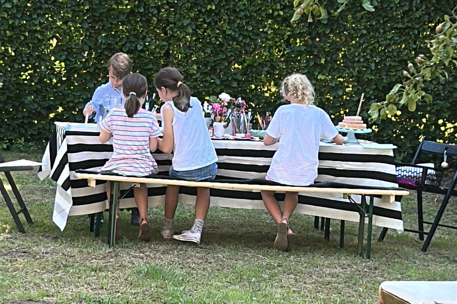 Vier Kinder sitzen von hinten an einem langen Gartentisch und gestalten gemeinsam