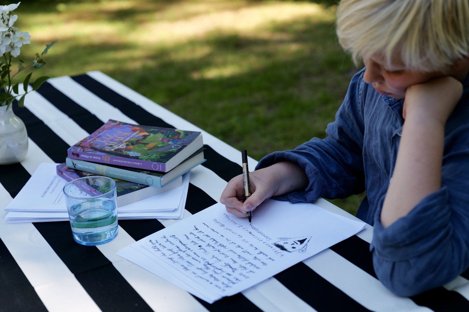 Ein Kind schreibt im Garten an einem Tisch, daneben die Bücher der Seldas-Haus-Trilogie